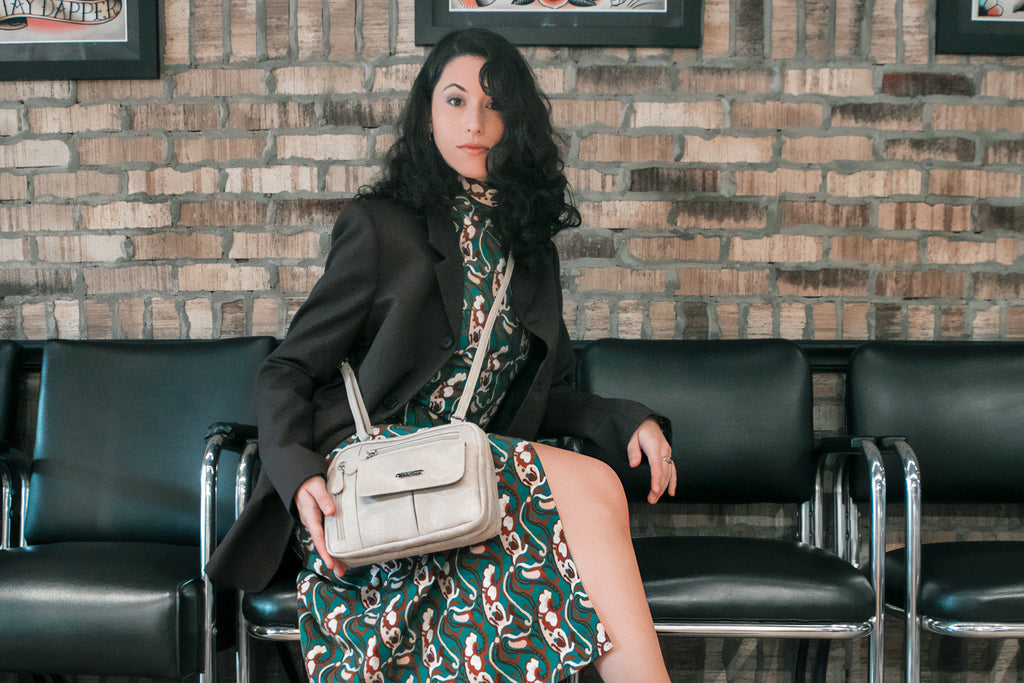 Best Bags for Moms with Toddlers - A woman in a black blazer with a green dress sits in a chair with her zippy triple compartment crossbody bag.
