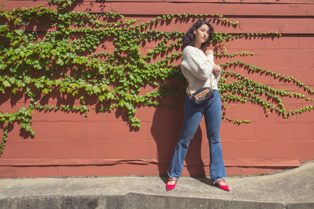 Affordable Gifts for Her - A woman in a white sweater, jeans, pink shoes, and a black and brown Delta Crossbody Bag poses in front of a brick wall covered by vines. 